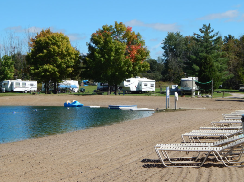 A sandy beach with lounge chairs, a calm lake, and RVs in the background under a clear blue sky.