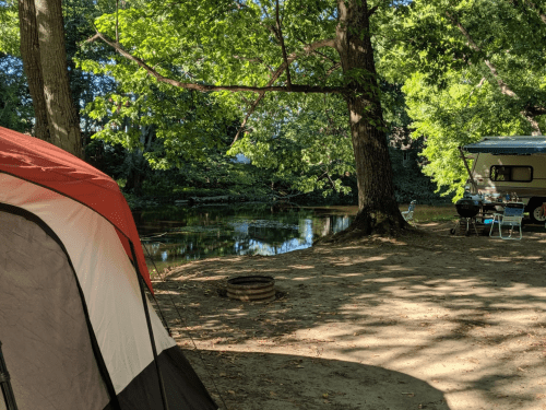 A campsite by a calm river, featuring a red and black tent, a camper, and trees providing shade.