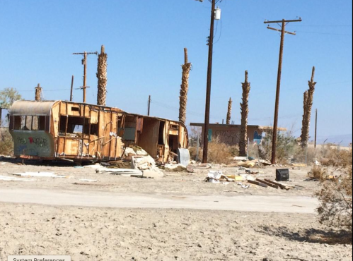 Abandoned trailer in a desolate landscape with dead palm trees and scattered debris under a clear blue sky.