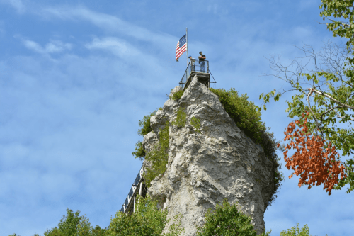 A person stands on a rocky outcrop with an American flag, surrounded by trees and a blue sky.