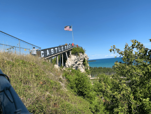 View of Castle Rock sign on a cliff with an American flag, overlooking a blue lake and green trees under a clear sky.