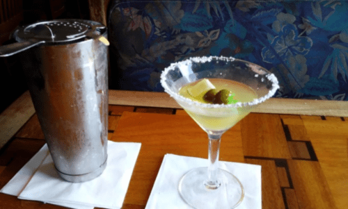 A martini glass with a lime and olive, next to a shaker on a wooden table with a patterned background.