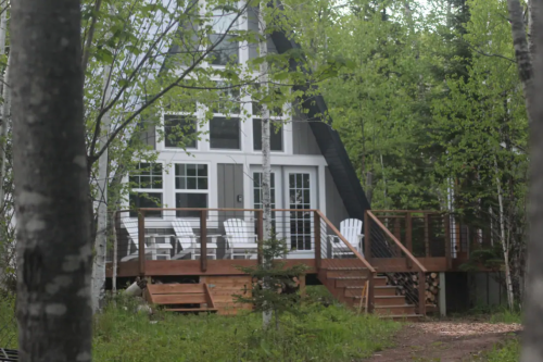 A modern A-frame cabin surrounded by trees, featuring a wooden deck with white chairs and a sloped roof.