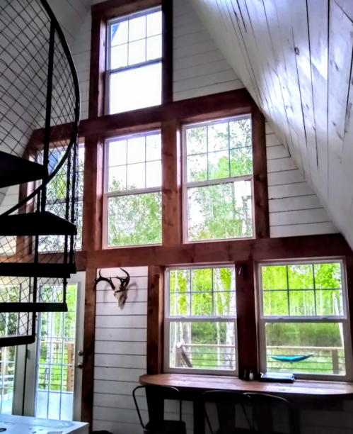 Interior of a cozy cabin with large windows, wooden beams, a spiral staircase, and a rustic table. Greenery outside.
