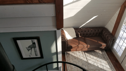 A cozy living room with a brown leather couch, framed artwork, and sunlight streaming through a window.