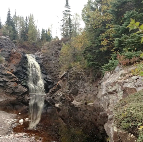 A serene waterfall cascades down rocky cliffs, surrounded by trees and autumn foliage, reflecting in a calm pool below.