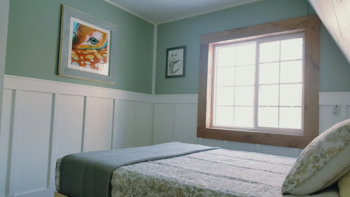 Cozy bedroom with a bed, green walls, framed artwork, and a large window letting in natural light.