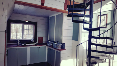 A cozy kitchen with a spiral staircase, featuring white walls, blue cabinets, and natural light from a window.