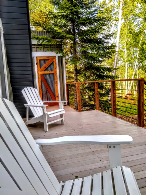 A wooden deck with two white chairs, surrounded by trees, featuring a red door in the background.
