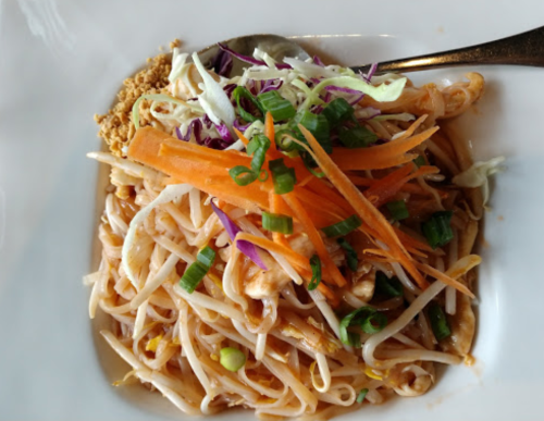 A plate of noodles topped with shredded carrots, cabbage, green onions, and crushed peanuts.