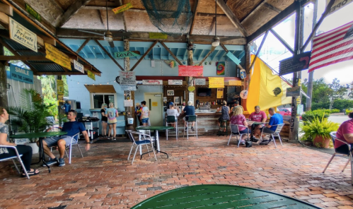 A lively outdoor dining area with patrons seated at tables, colorful signs, and a bar in a tropical setting.