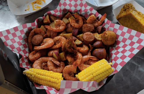 A basket of shrimp, sausage, potatoes, and corn on the cob, served with a checkered paper lining.