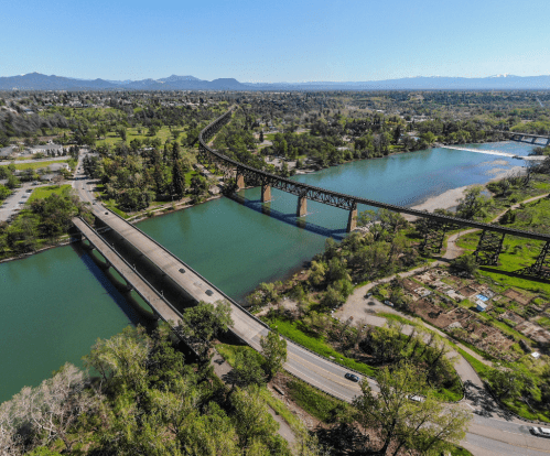 Aerial view of a river with bridges, surrounded by greenery and mountains in the background on a clear day.