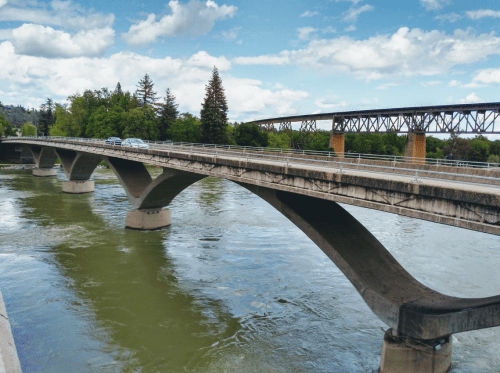 A concrete bridge spans a river, with trees and a railway bridge visible in the background under a partly cloudy sky.