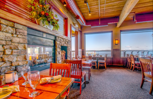 Cozy restaurant interior with wooden tables, chairs, a stone fireplace, and large windows overlooking a scenic view.