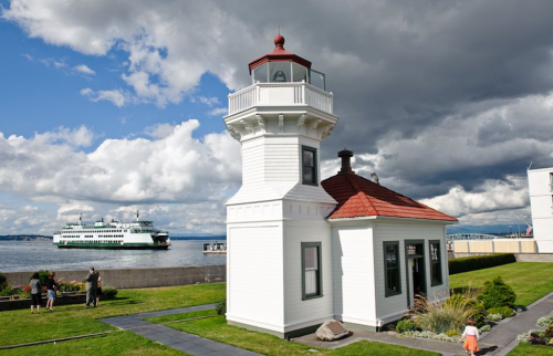 A white lighthouse with a red roof beside a waterfront, with a ferry passing in the background under a cloudy sky.