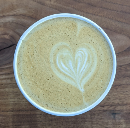 A close-up of a latte in a white cup, featuring a heart-shaped foam design on top, set on a wooden surface.
