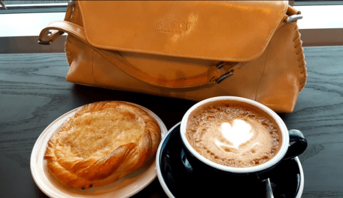 A pastry on a plate next to a cup of coffee with a heart-shaped foam design, beside a stylish tan handbag.