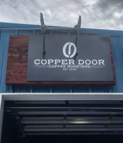 Sign for Copper Door Coffee Roasters, established in 2008, displayed on a blue building under a cloudy sky.