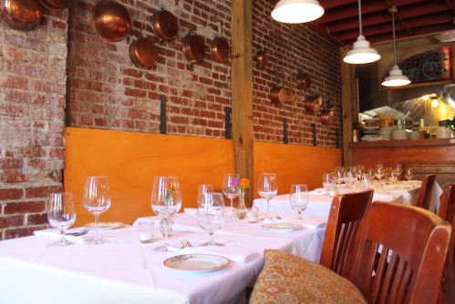 A cozy restaurant interior with wooden tables set for dining, featuring exposed brick walls and copper pots hanging.