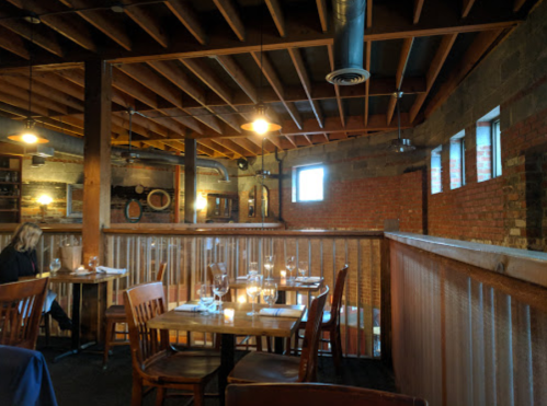 Cozy restaurant interior with wooden tables, chairs, and exposed brick walls, featuring warm lighting and a rustic atmosphere.
