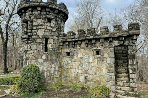 A weathered stone castle with a turret, surrounded by bare trees and greenery, set against a cloudy sky.