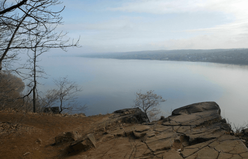 A serene view of a calm river surrounded by trees and rocky terrain under a cloudy sky.