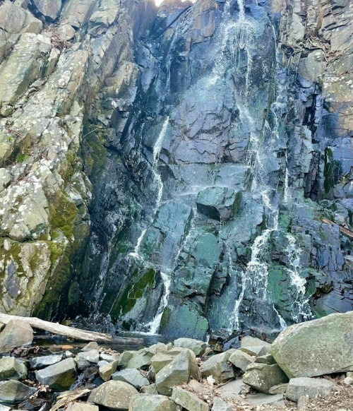 A cascading waterfall flows down a rocky cliff, surrounded by moss-covered stones and a serene natural setting.