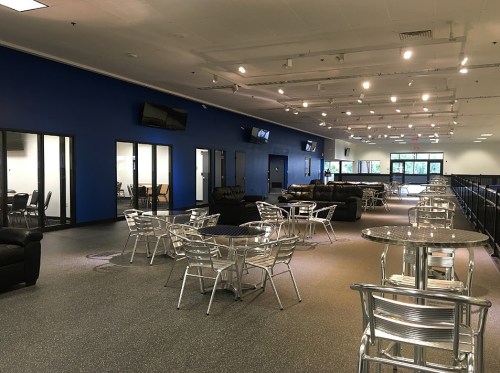 A spacious indoor area with blue walls, tables, chairs, and seating areas, featuring multiple TVs and large windows.