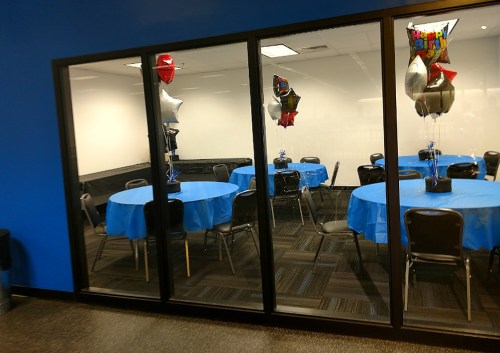 A party room with blue tablecloths, chairs, and balloons, viewed through glass windows.