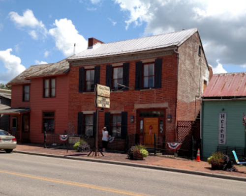 Historic brick building with a sign, surrounded by colorful houses and decorative plants, under a partly cloudy sky.