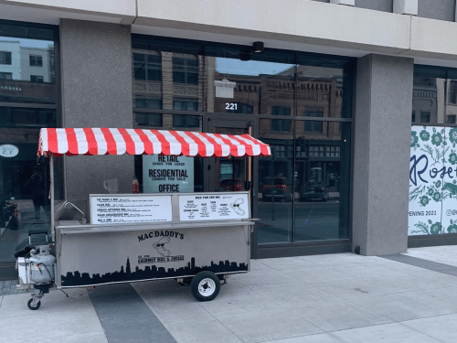 A food cart with a red and white striped awning parked outside a building with large windows and a "For Rent" sign.