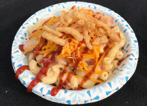 A paper plate filled with macaroni and cheese topped with shredded cheese, crispy onions, and barbecue sauce.