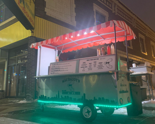 A food cart named "Mac Daddy's" with a red and white striped canopy, illuminated at night on a city street.