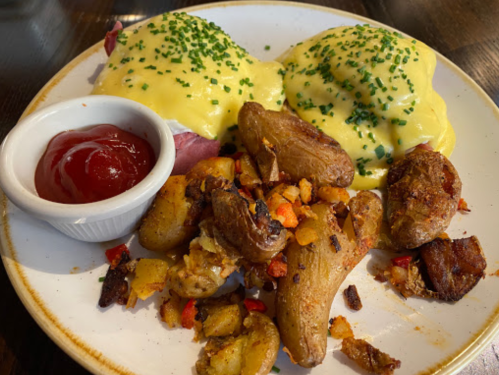 Plate of eggs Benedict with hollandaise sauce, chives, roasted potatoes, and a side of ketchup.