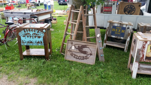 A collection of rustic wooden signs and furniture for sale, including "Welcome" signs and a ladder display, set in a grassy area.