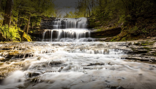 A serene waterfall cascades over rocky steps, surrounded by lush green trees and a tranquil atmosphere.