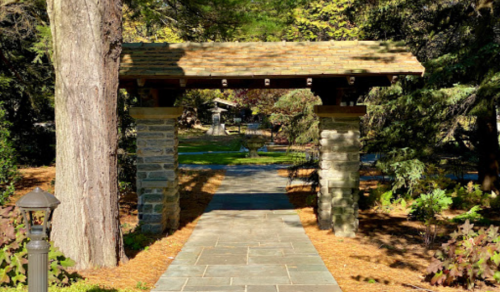 A stone archway with a wooden roof frames a pathway leading through a lush, green garden.