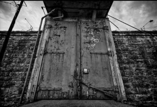 A pair of large, weathered metal doors set against a stone wall, under a cloudy sky.