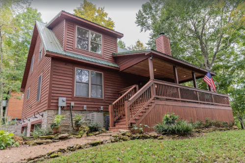 A red wooden house with a porch, surrounded by greenery and featuring an American flag.