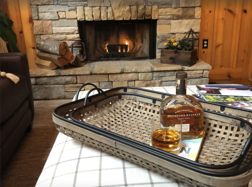 A cozy living room scene featuring a stone fireplace, a basket with a whiskey bottle, and a magazine on a table.