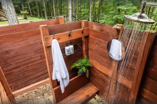Outdoor shower with wooden walls, a showerhead, towels, toiletries, and a mirror, surrounded by greenery.