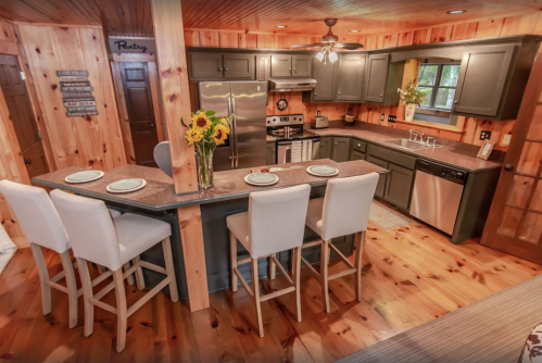 Cozy kitchen with wooden walls, gray cabinets, stainless steel appliances, and a bar with four stools and sunflowers.
