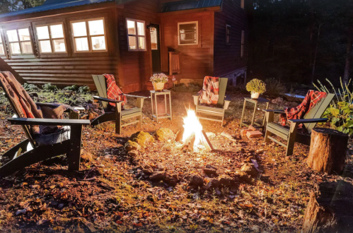 A cozy outdoor campfire setup with chairs and blankets, surrounded by trees and a cabin in the background.