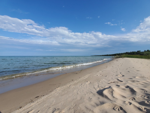 A serene beach scene with gentle waves, soft sand, and a clear blue sky with scattered clouds.