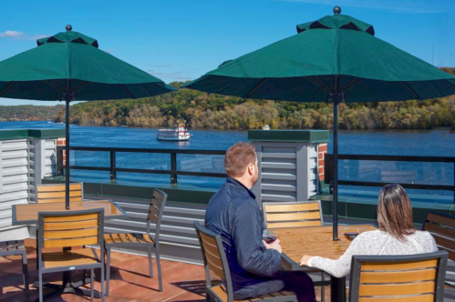 A couple sits at a table on a rooftop patio, overlooking a river with a boat and trees in the background.