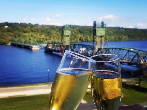 Two glasses of champagne clink together, with a scenic river and bridge in the background.