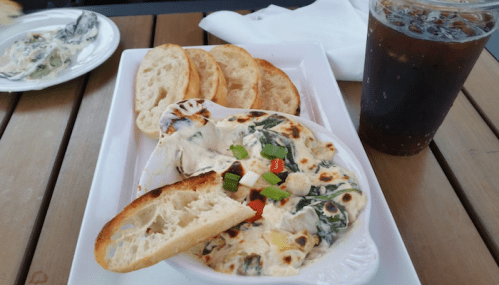 A creamy spinach dip served in a dish with toasted bread slices and a glass of iced drink on a wooden table.