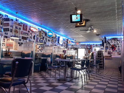 A retro diner interior with blue lighting, checkered floor, and walls adorned with vintage photos and memorabilia.