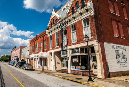 Historic brick buildings line a street, featuring shops and a theater under a blue sky with fluffy clouds.